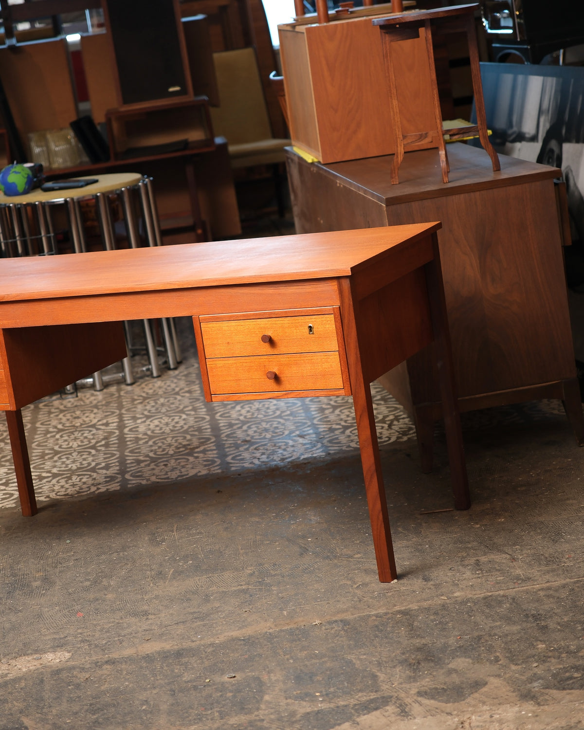 Mid-century modern Danish teak desk shown in a warehouse setting beside other vintage furniture.