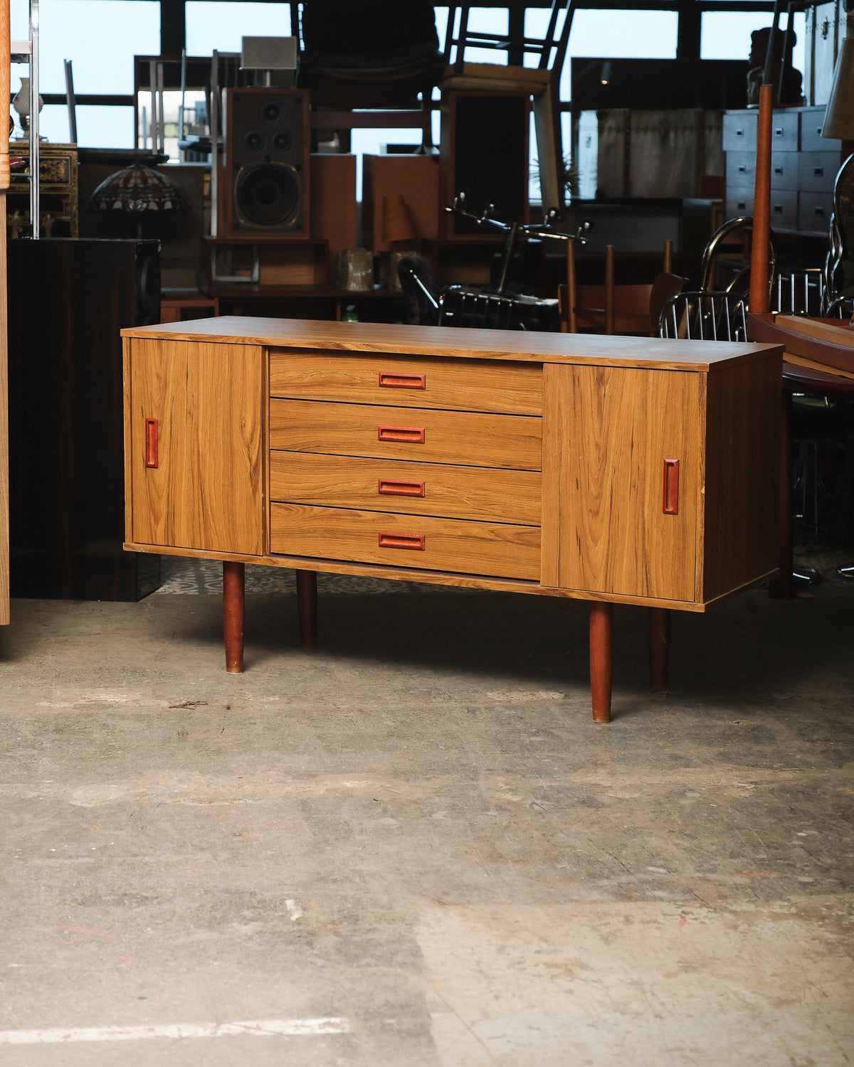 Mid-century modern teak sideboard with sliding doors and four drawers, shown in a warehouse setting.