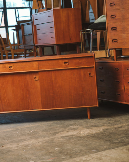A mid-century modern wooden sideboard or credenza with warm orange-brown wood grain, shown in a showroom or warehouse setting with other furniture pieces in the background.
