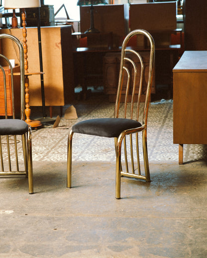 Two brass dining chairs shown in a showroom setting alongside other vintage furniture.