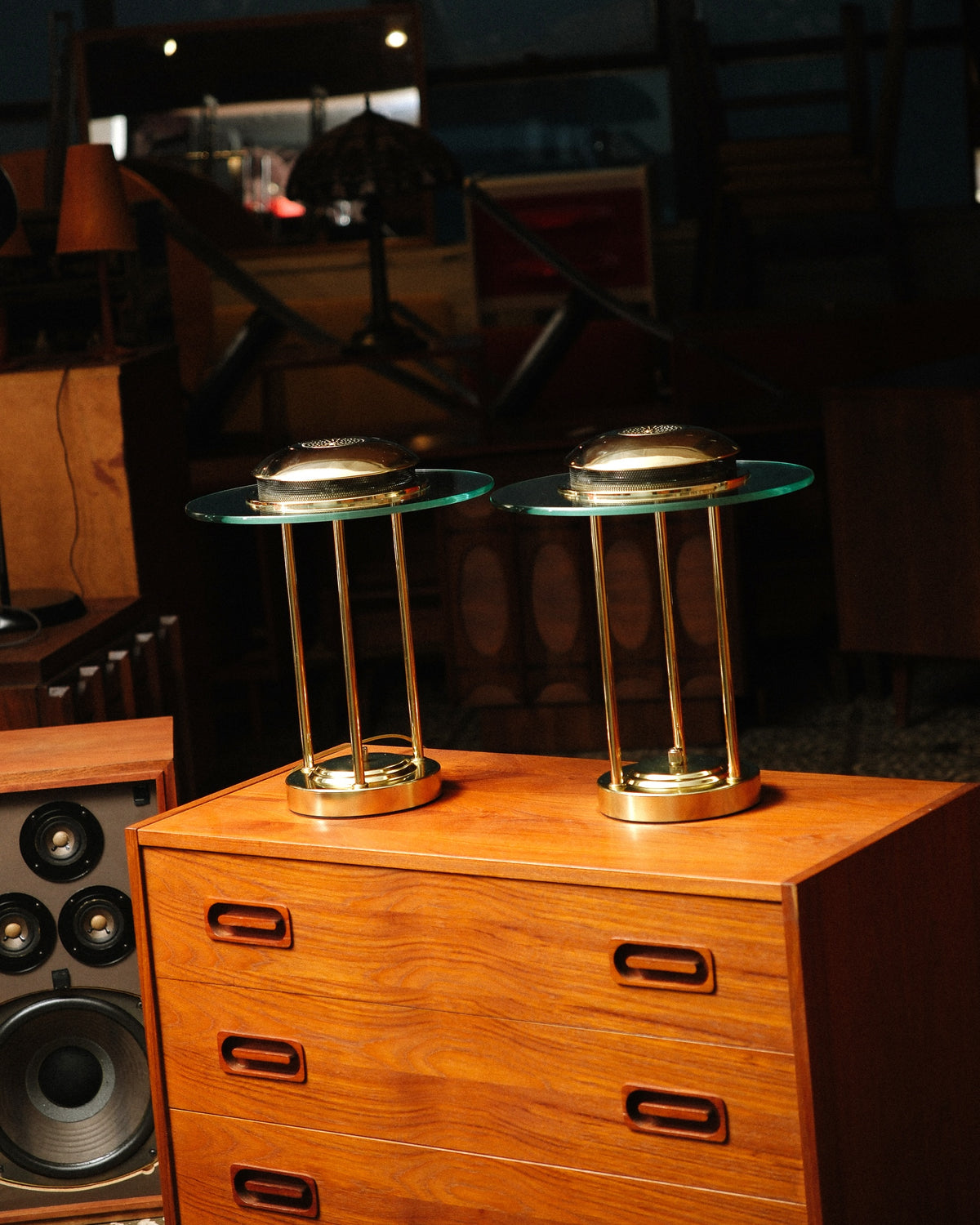 Two matching brass saturn lamps side by side on a teak dresser in a showroom setting.