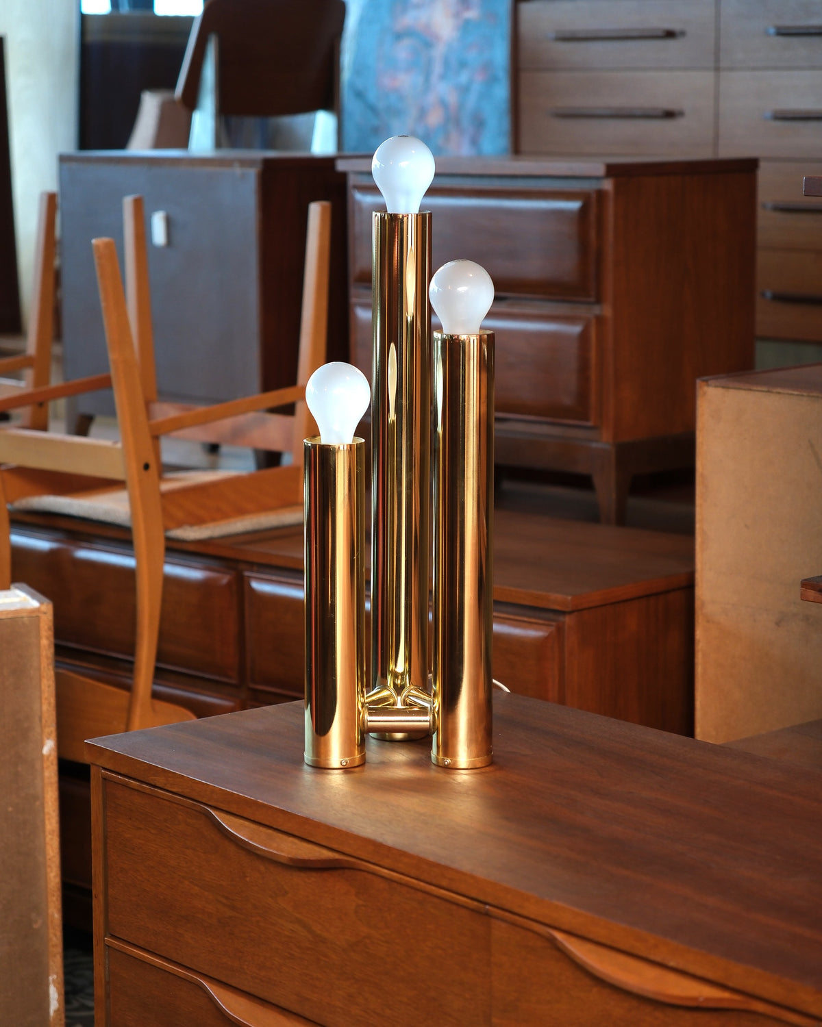 Vintage brass table lamp with three staggered tubes displayed on a dresser in a furniture showroom