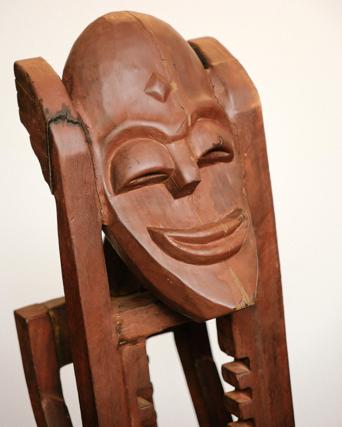 Close-up of the carved wooden mask face with closed eyes and a smiling expression, showing natural wood grain and a crack.