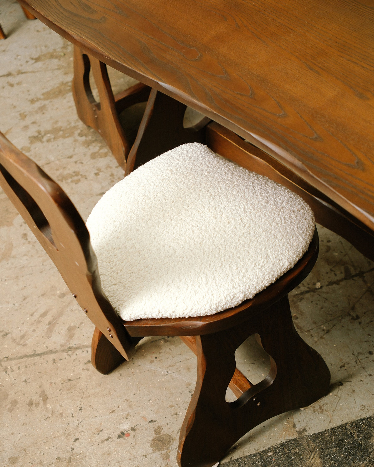 Boucle-upholstered chair seat tucked under the oak dining table, showing sculptural cut-out legs.