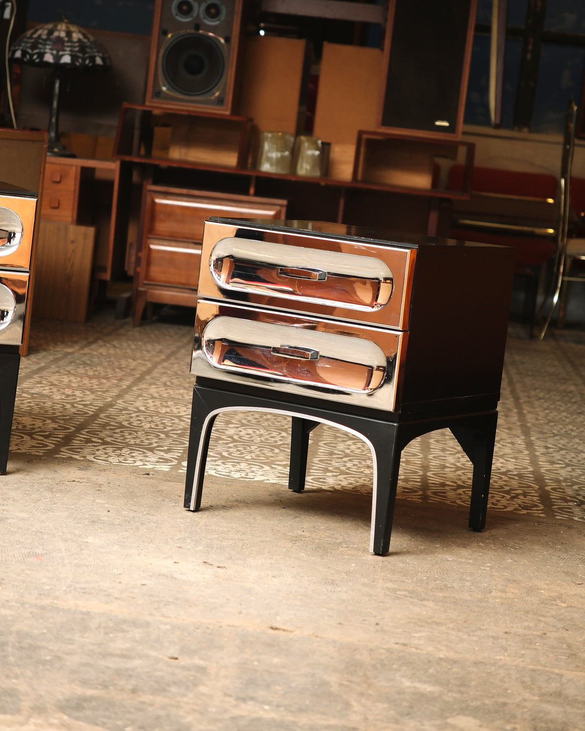 Single chrome bedside table with two drawers, shown against showroom backdrop.