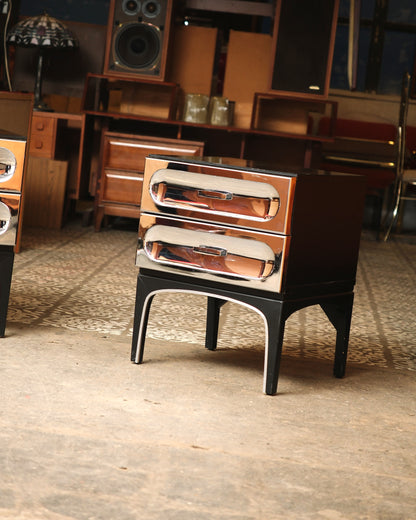 Single chrome bedside table with two drawers, shown against showroom backdrop.