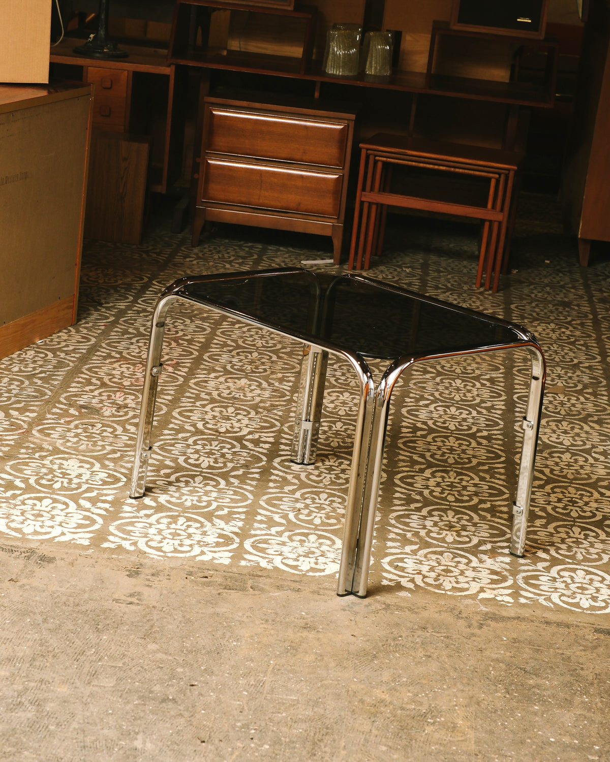 Vintage chrome coffee table with smoked glass top shown in a showroom setting on patterned floor tile.