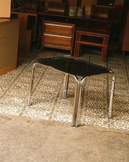Vintage chrome coffee table with smoked glass top shown in a showroom setting on patterned floor tile.