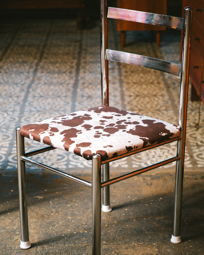 Close angled view of a single chrome chair showing ladder-back frame and cowhide print seat.