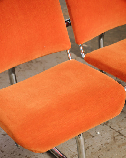 Close-up of two orange tubular chairs showing the velvet upholstery and chrome frame joints.