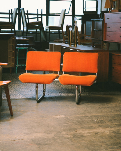 Front view of two orange tubular chairs against a backdrop of teak dressers and lamps.