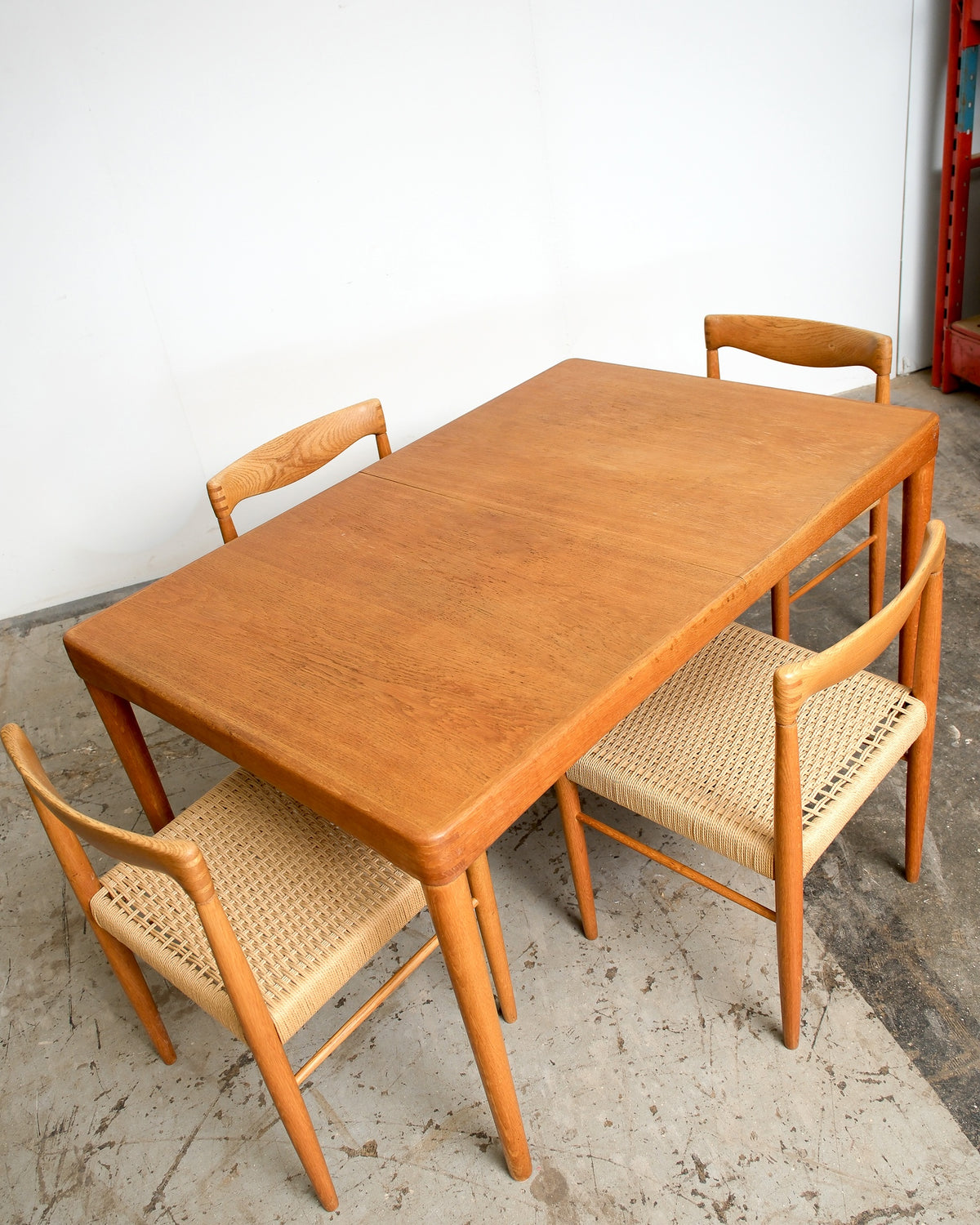 Full view of the oak dining table with four cord-seat chairs arranged around it, in a warehouse setting.