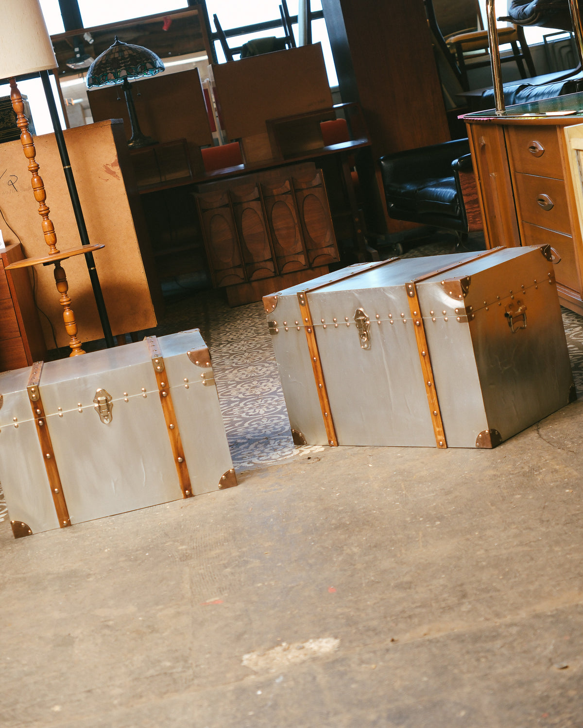 Both trunks shown side by side in the showroom among other vintage furniture pieces.