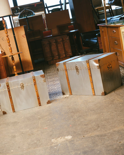 Both trunks shown side by side in the showroom among other vintage furniture pieces.