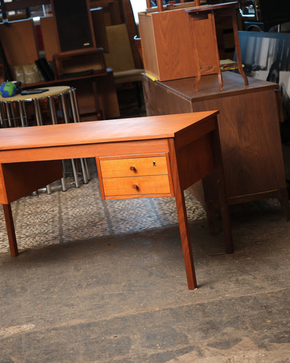 Mid-century modern Danish teak desk shown in a warehouse setting beside other vintage furniture.