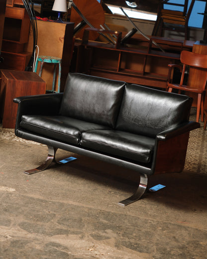 Angled view of the black leather sofa showing curved wood side panel and flared chrome legs.