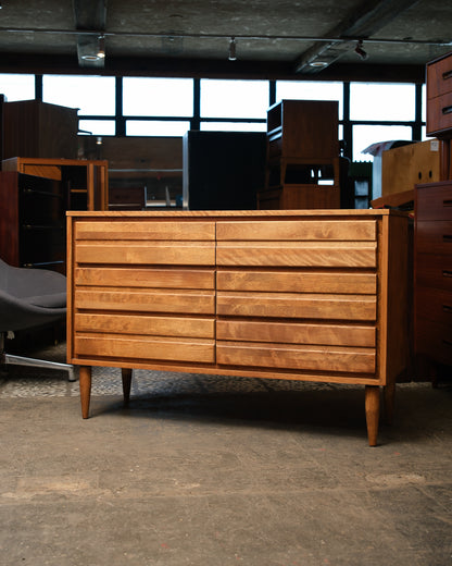 Wooden dresser in a storage or warehouse setting with other furniture in the background.