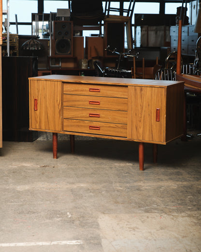 Mid-century modern teak sideboard with sliding doors and four drawers, shown in a warehouse setting.