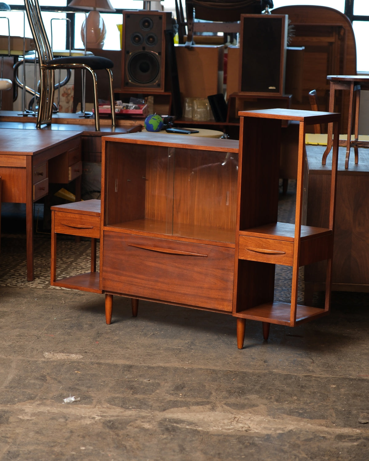 Mid-century modern walnut display cabinet with sliding glass door and asymmetrical open shelving, shown in warehouse setting.