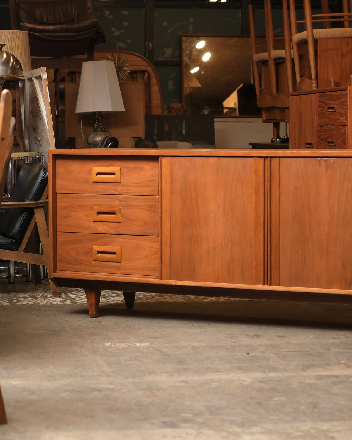 Side view of mid-century modern walnut dresser sideboard showing three drawers and sliding cabinet doors, shown among