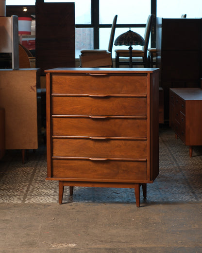 Mid-century modern walnut tallboy dresser with five drawers and sculpted pulls, shown straight on in a warehouse setting.