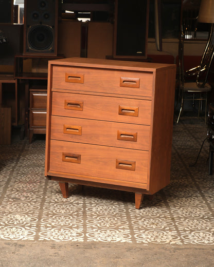 Mid-century walnut 4-drawer tallboy dresser shown at an angle with tapered legs and sculpted pulls, in a warehouse setting.