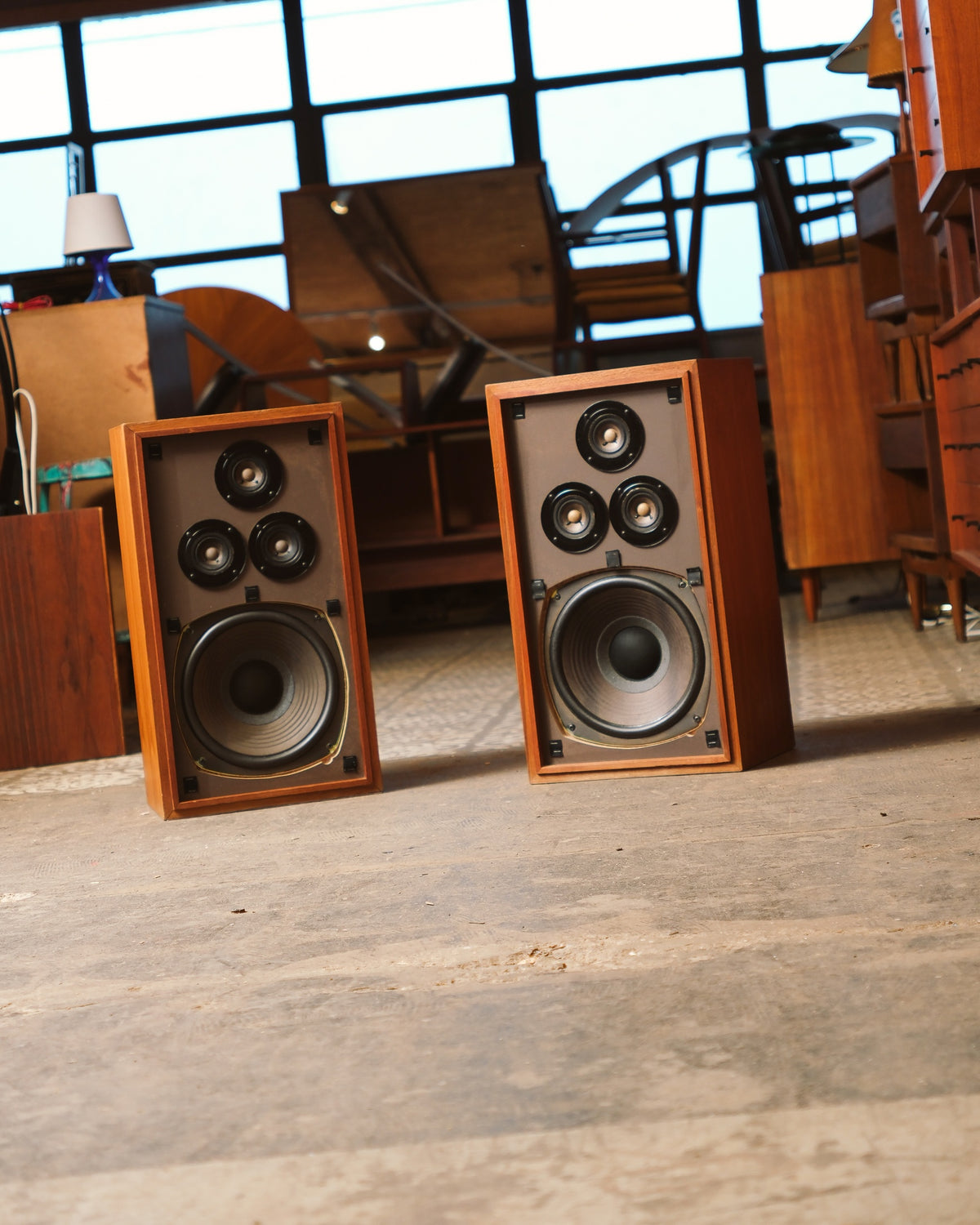 Angled view of the two speakers standing on a patterned floor, showroom furniture in the background.