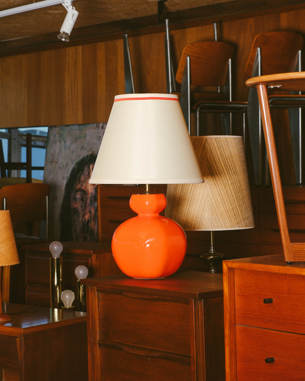 Orange table lamp displayed on a wood dresser in a showroom setting with other vintage furniture.