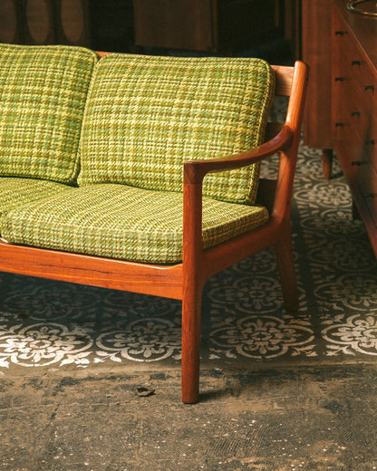 Side angle of the sofa showing the teak arm, leg, and green plaid cushions.