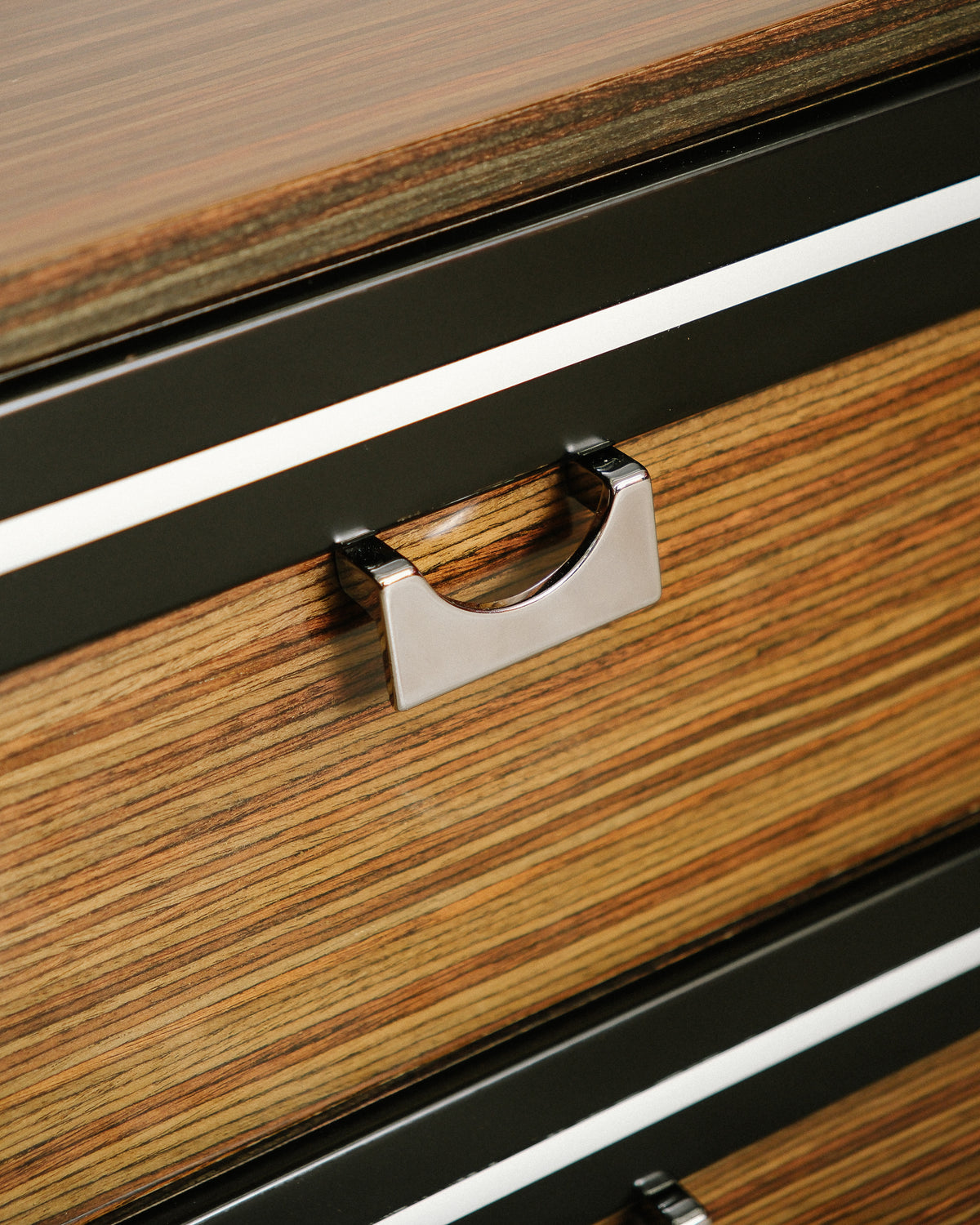 Close-up detail of the chrome pull and black-and-white trim between drawers on zebrawood front.